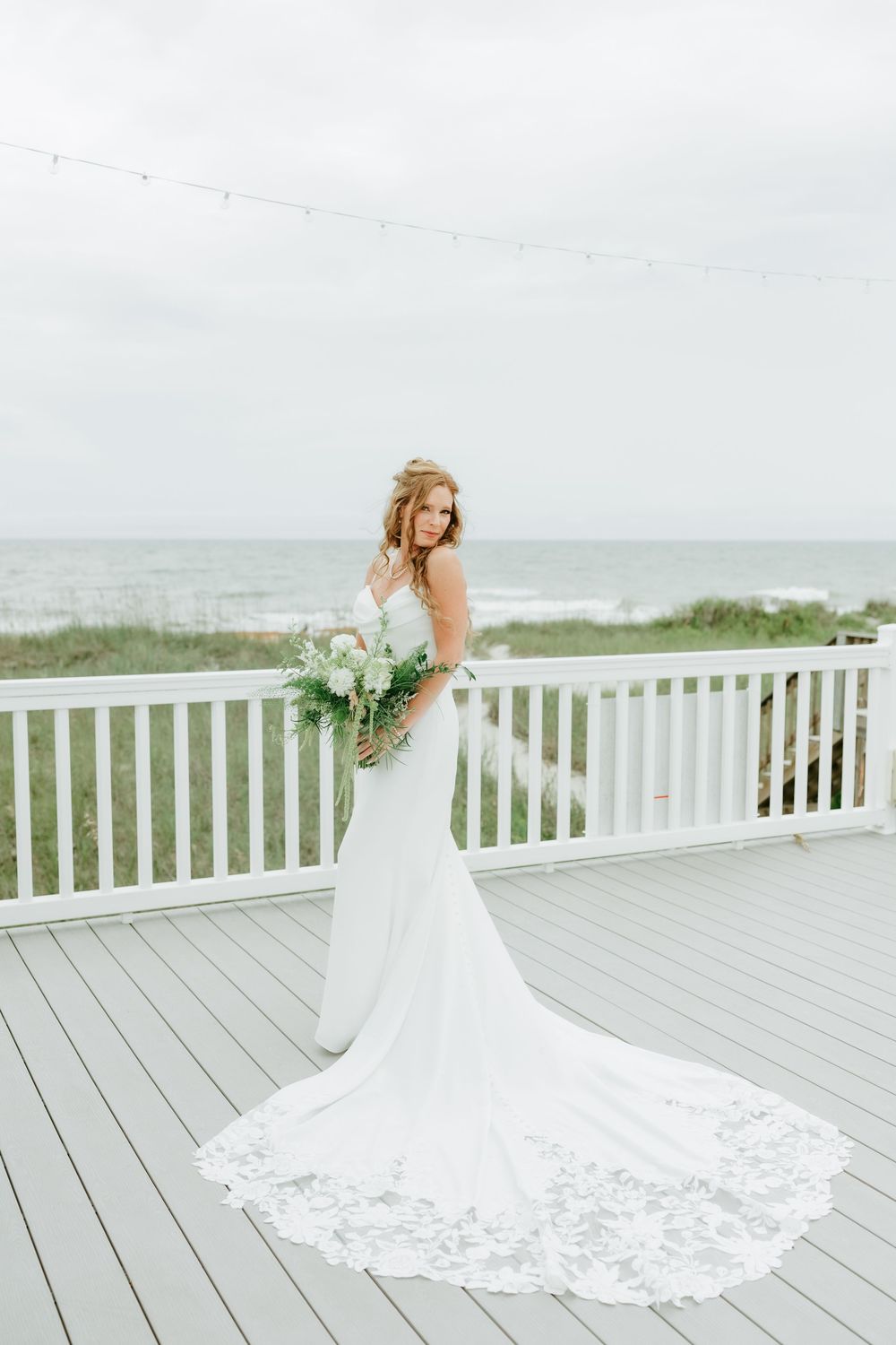 Bride in white lace mermaid gown holding bouquet on beachfront deck with ocean views.