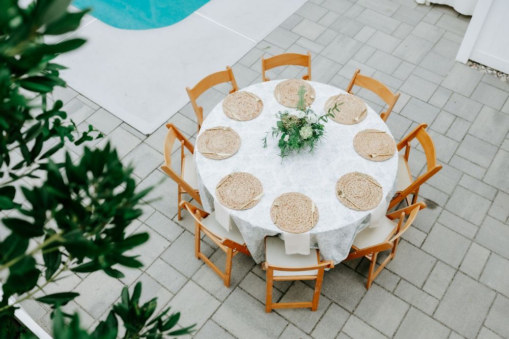 Elegant outdoor round table with white linen, wicker placemats, wooden chairs, and floral centerpiece near a pool.