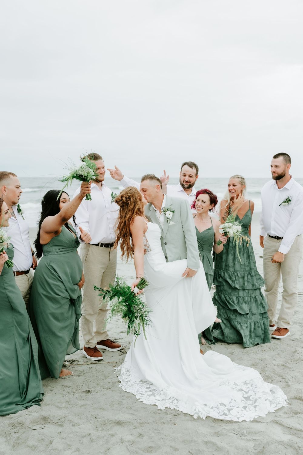 Wedding party celebrates on beach, bride and groom kiss surrounded by groomsmen in tan and bridesmaids in sage green gowns.