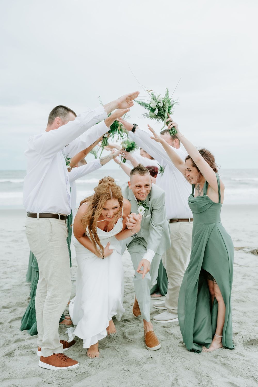 Wedding party celebrating on beach, bride and groom ducking under arch of raised hands holding bouquets, bridesmaids in sage green.