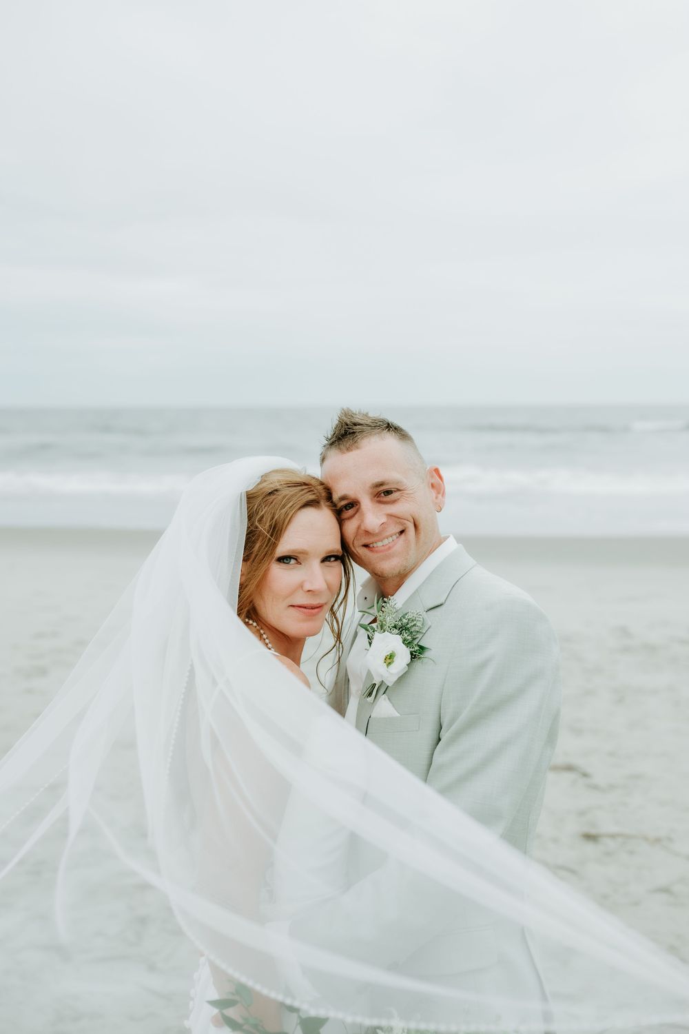 A bride and groom pose romantically on a beach, her veil flowing in the wind, both smiling in wedding attire.