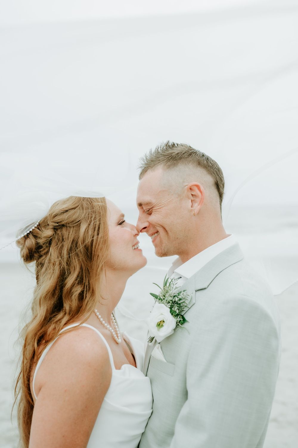 A bride and groom share a romantic nose-to-nose moment outdoors, dressed in white and light gray wedding attire.