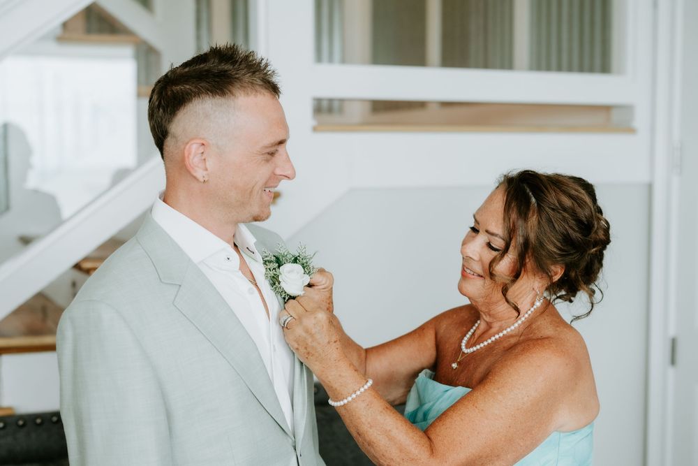A woman in a blue dress pins a white boutonniere on a smiling groom in a light gray suit at a wedding.