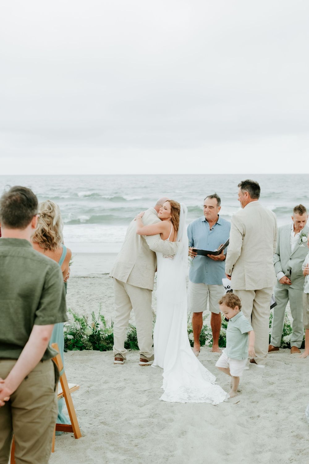 Bride and groom share first kiss during romantic beach wedding ceremony with ocean waves and wedding party behind them.