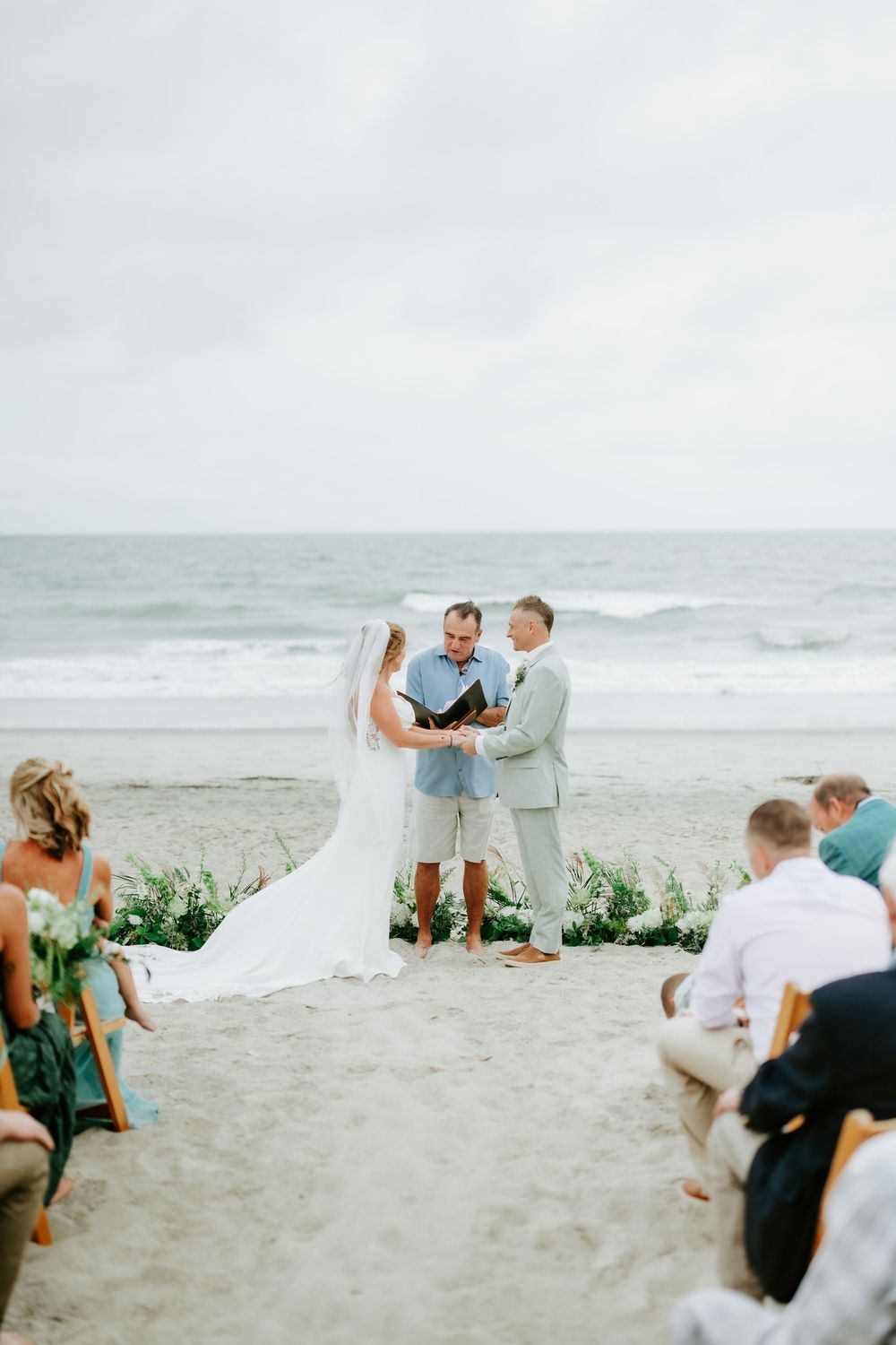Romantic beach wedding ceremony with bride and groom exchanging vows by the ocean, officiant holding book, guests seated on sand.