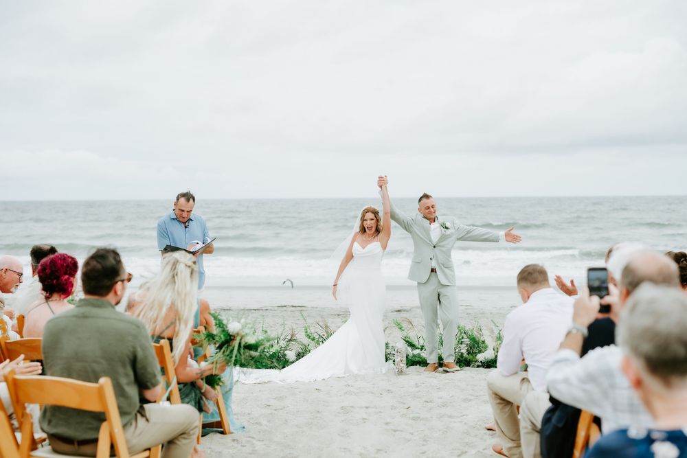 Joyful newlyweds celebrate at a beach wedding ceremony with guests seated on wooden chairs by the ocean.