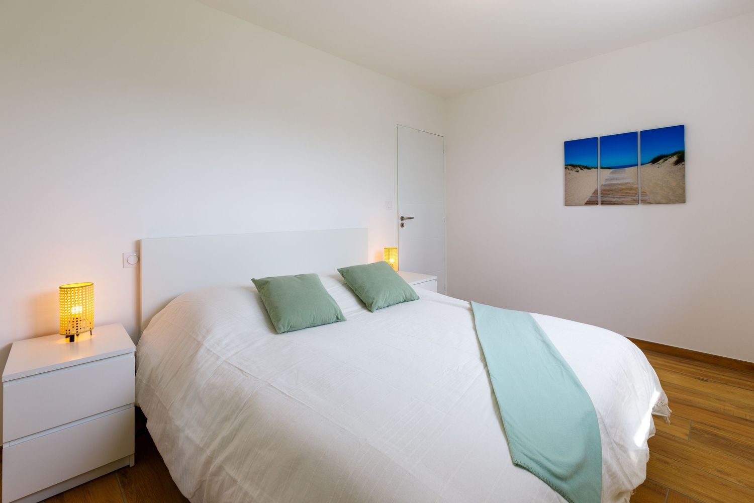 Chambre lumineuse aux tons blancs avec un lit double, des coussins d'appoint verts, une table de chevet en bois avec une lampe