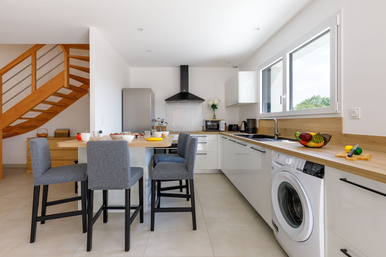 Cuisine ouverte avec des placards blancs, des plans de travail en bois, un coin repas, un escalier et un lave-linge intégré.