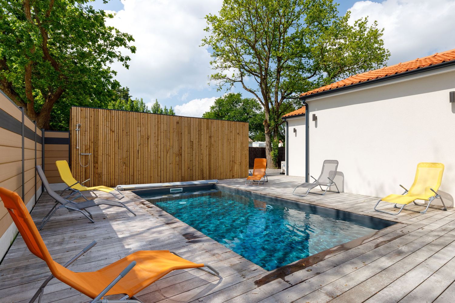 Piscine extérieure avec terrasse en bois, chaises longues colorées et clôture en bois, dans un jardin privé.