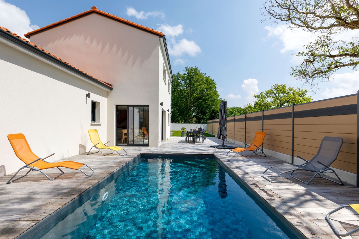 Villa moderne avec piscine rectangulaire, chaises longues orange et terrasse en bois sous un ciel bleu.