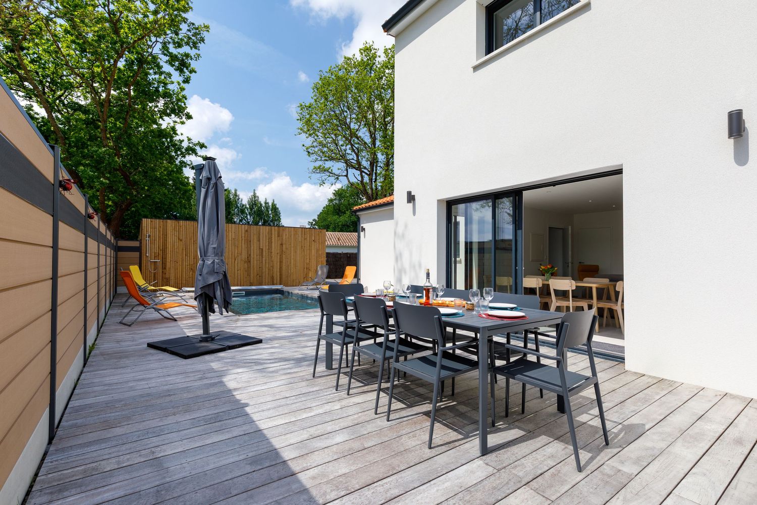 Terrasse extérieure  en bois au bord de la piscine, parasol et portes vitrées ouvertes