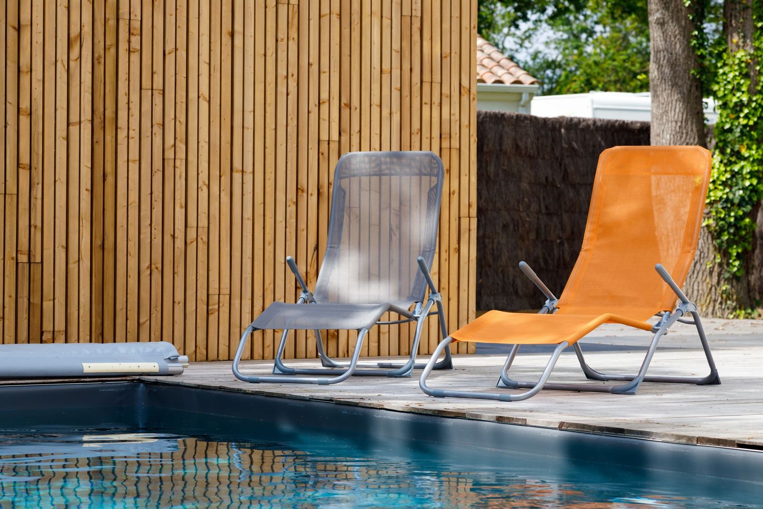 Deux chaises longues au bord d'une piscine, l'une grise et l'autre orange, disposées devant une clôture en bambou par une journée ensoleillé