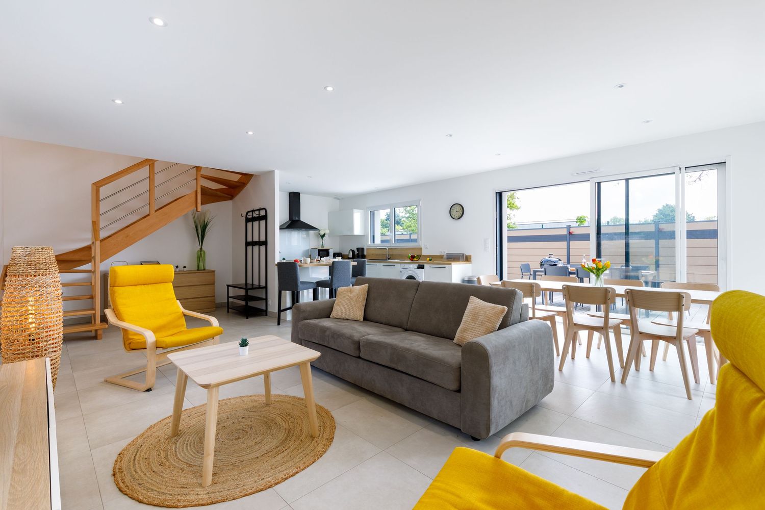 Salon avec canapé gris, chaises jaunes, escalier en bois et coin repas doté de grandes fenêtres.