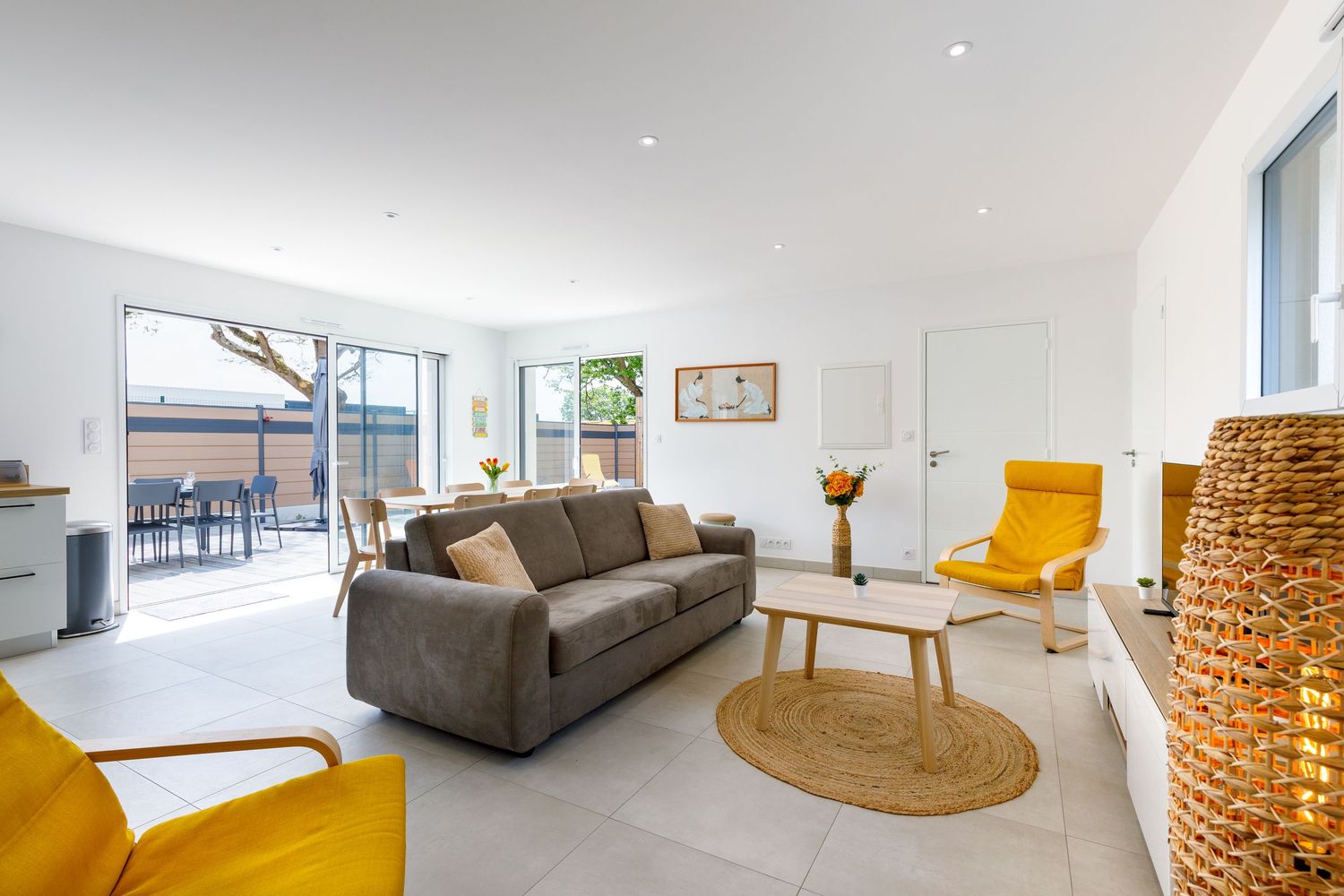 Salon grand lumineux avec un canapé gris, des fauteuils jaunes, une décoration en osier et de grandes baies vitrées donnant sur une terrasse