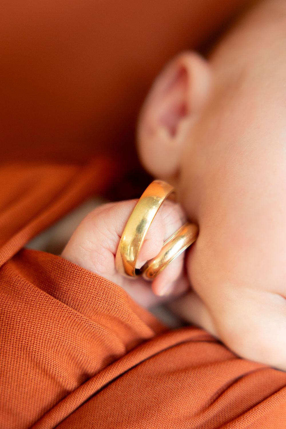 Close-up of newborn baby hand holding two gold wedding rings on orange fabric.