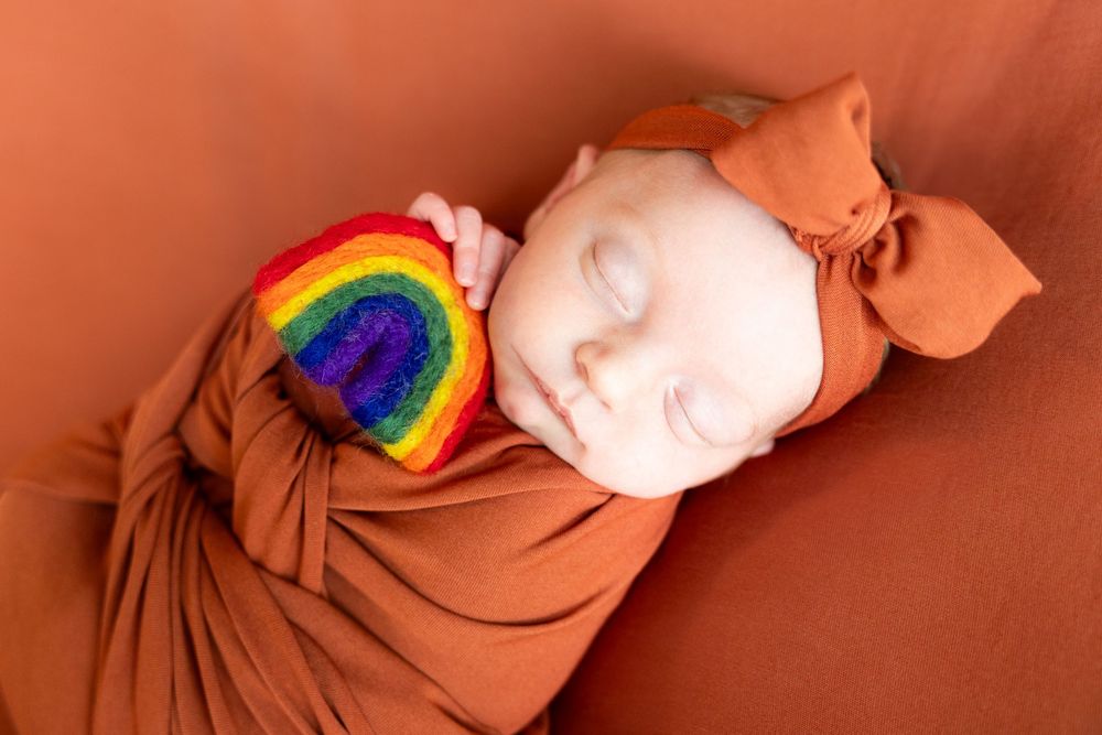 Sleeping newborn baby swaddled in rust orange wrap holding a colorful rainbow toy.