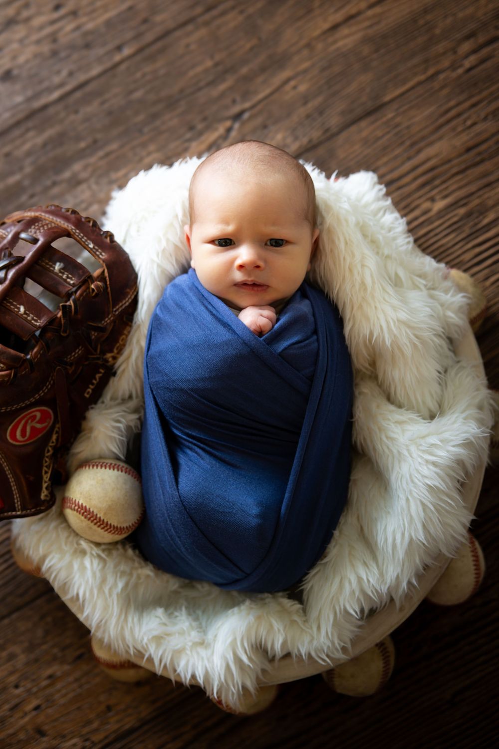 Alert newborn baby wrapped in blue swaddle posed in fur bowl with baseball glove and ball.