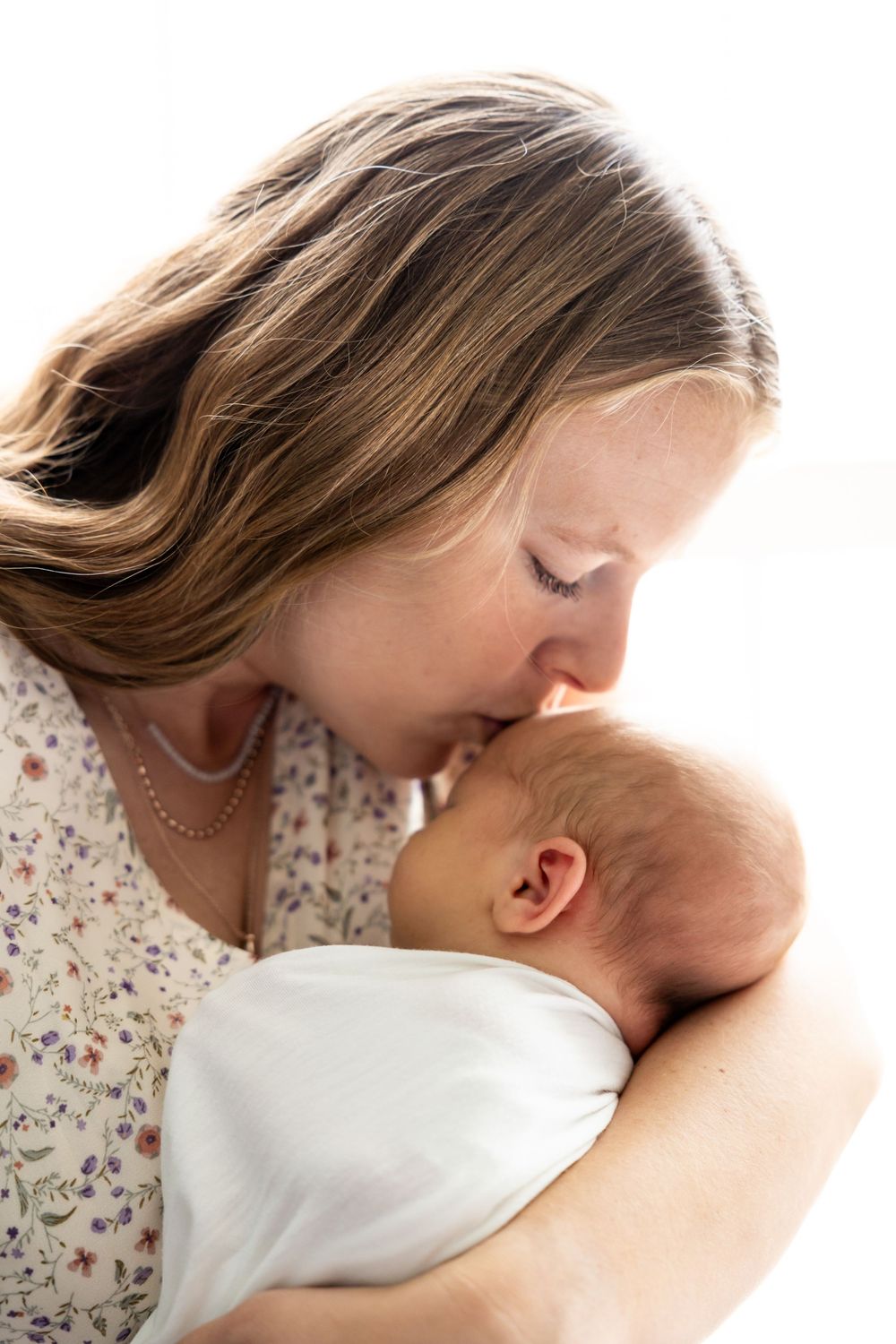 A mother tenderly kisses her swaddled newborn baby on the forehead against a white background.