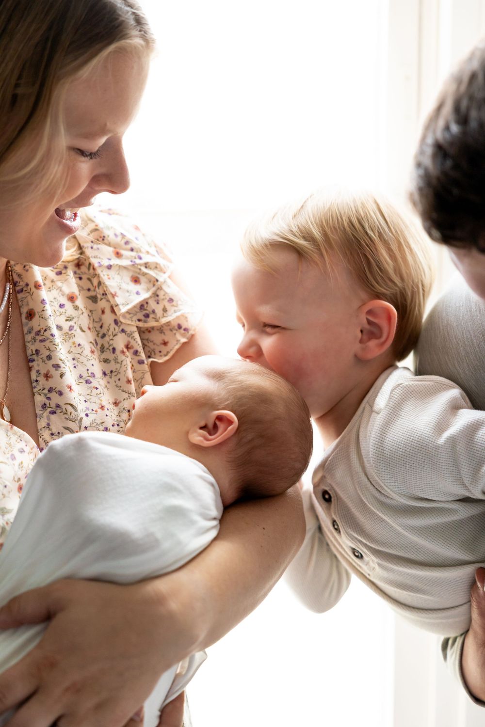 Mother smiling at newborn baby while toddler sibling leans in to give a gentle kiss.