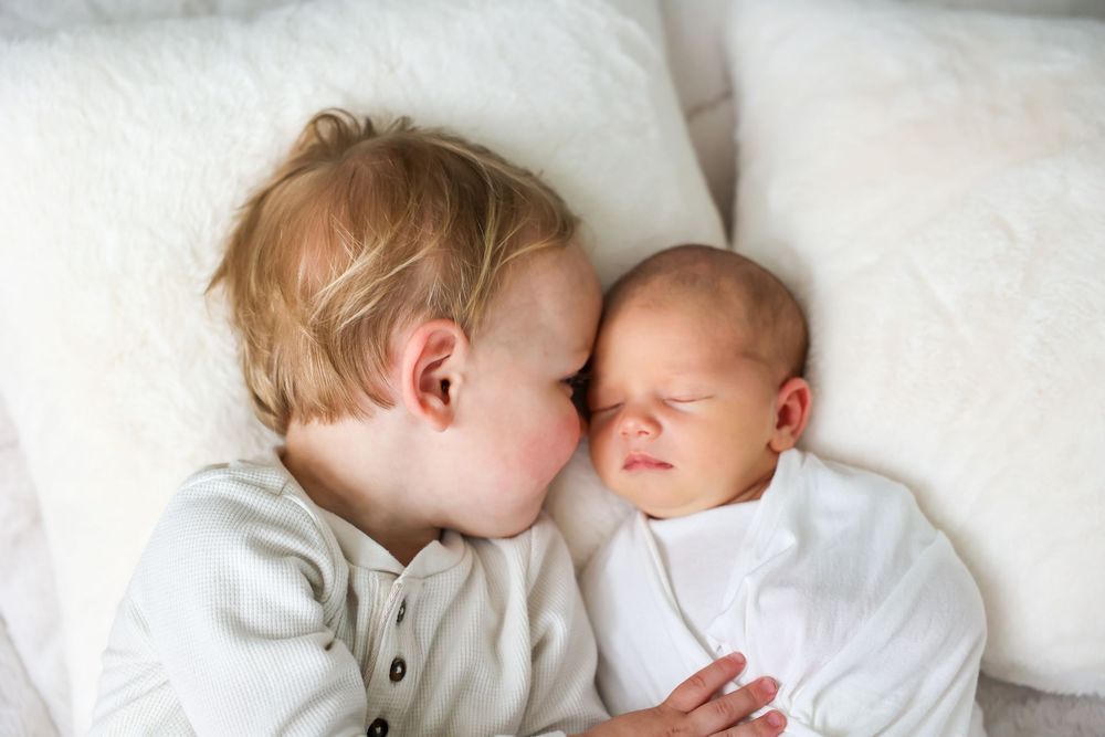 Toddler boy gently kissing sleeping newborn baby sibling lying on white bedding.