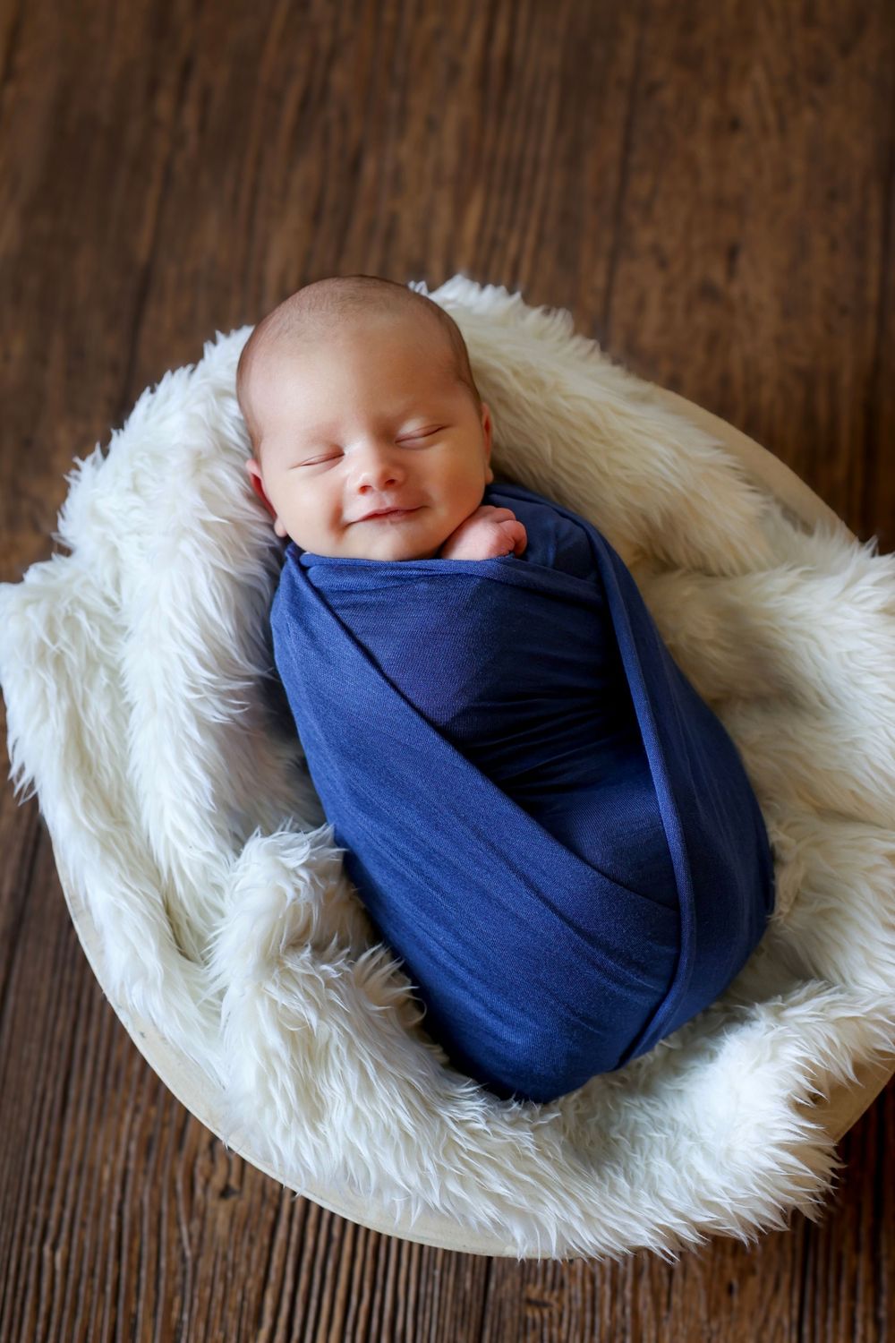 Smiling newborn baby swaddled in a blue wrap nestled in a white fur-lined wooden bowl.