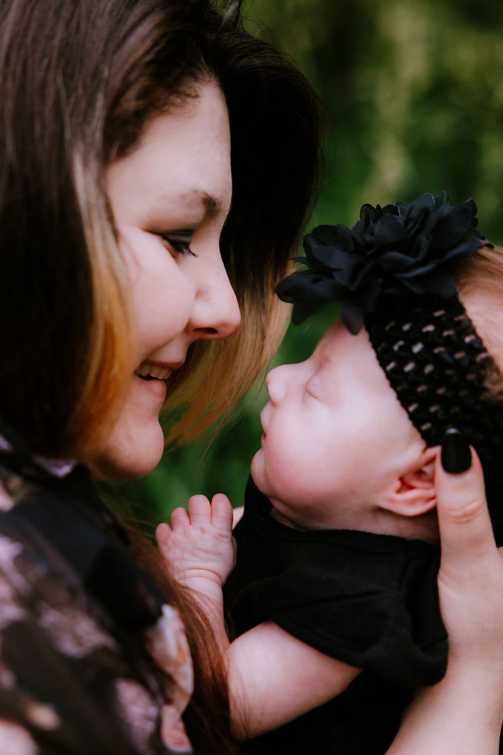 A smiling mother gazes lovingly at her newborn baby girl wearing a black floral headband outdoors.