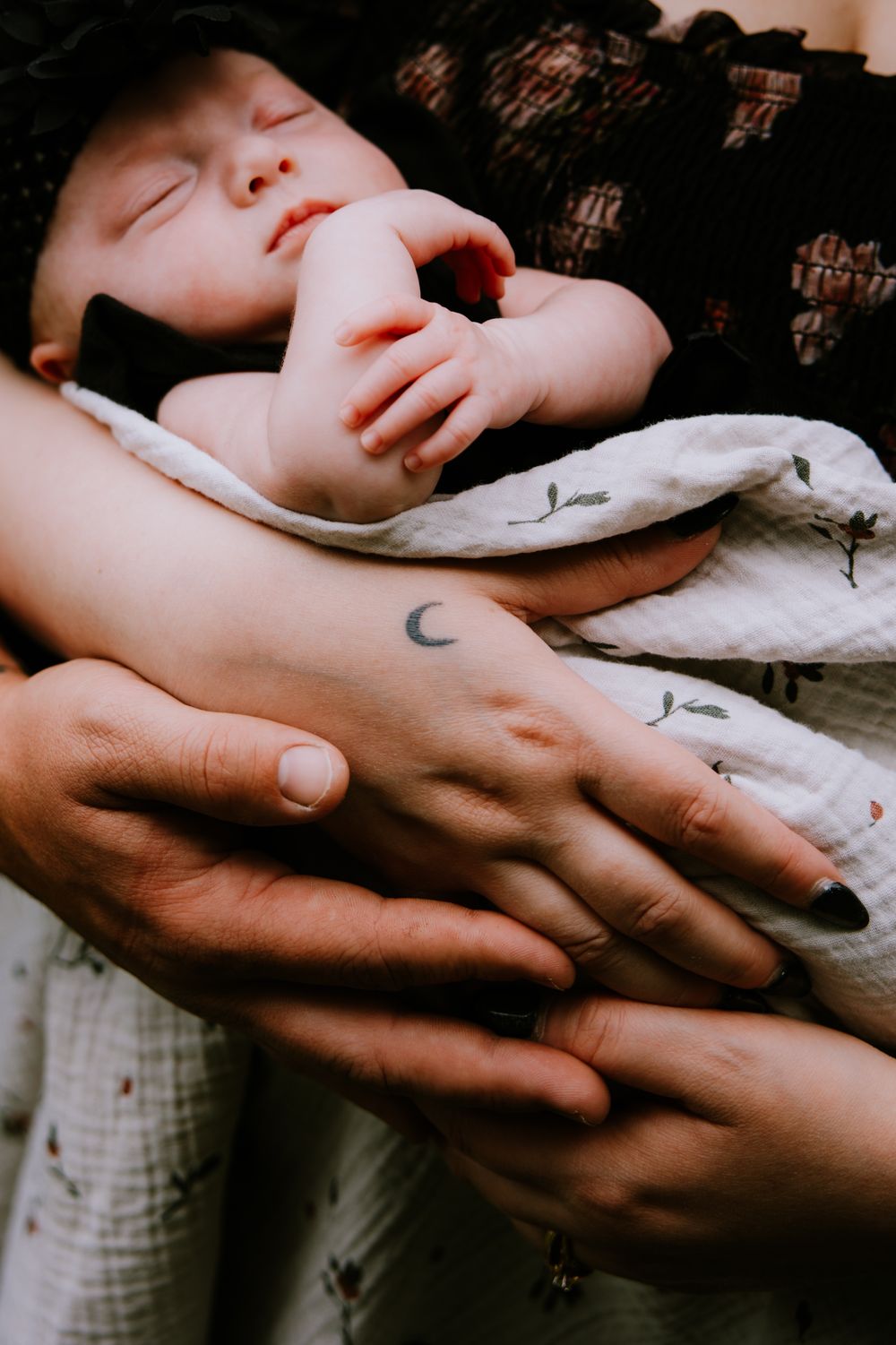 Sleeping newborn baby cradled in parents hands, with a small crescent moon tattoo visible on wrist.