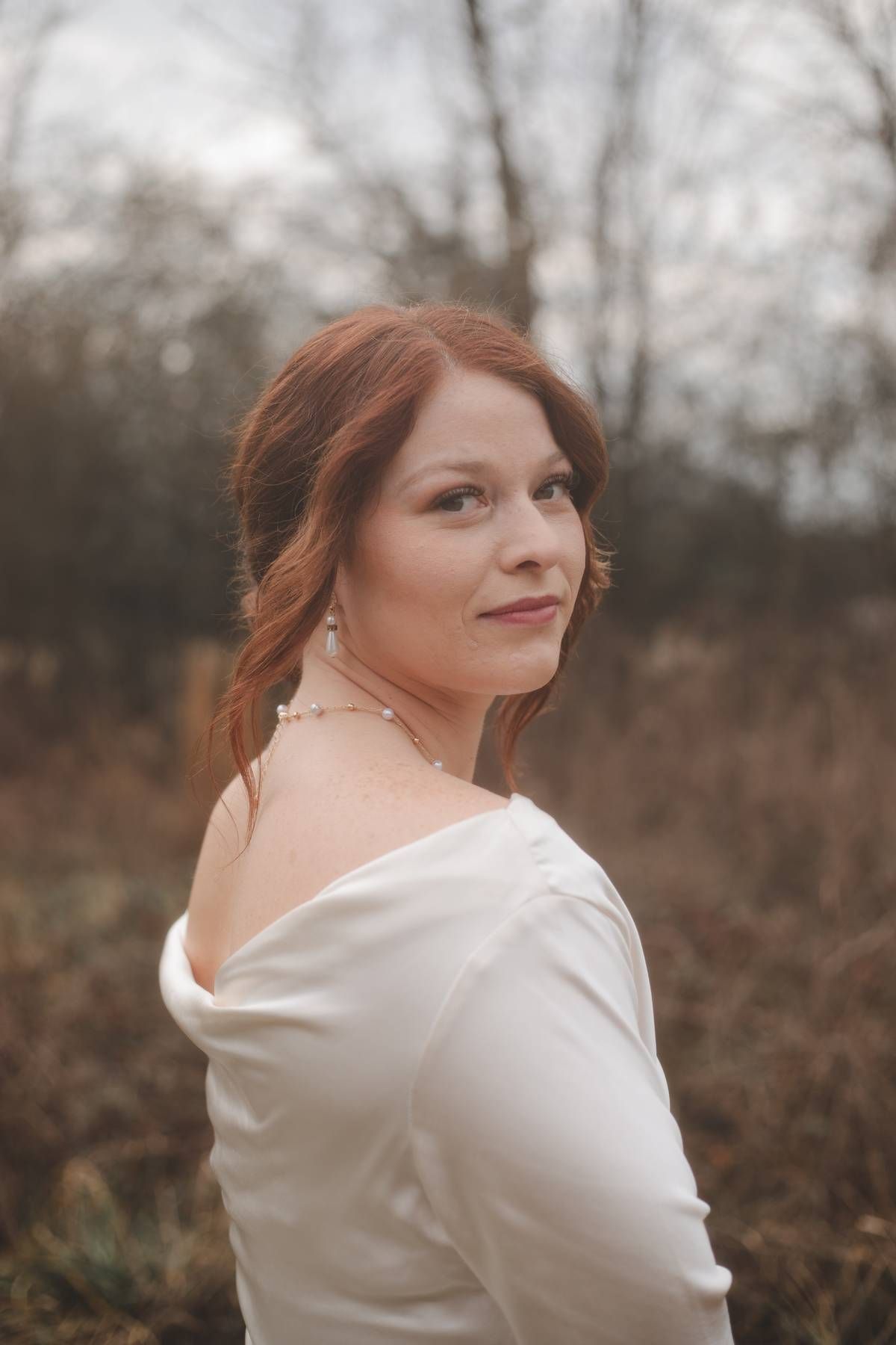 A red-haired woman in a cream satin top looks over her shoulder with a soft smile in a moody autumn field portrait.