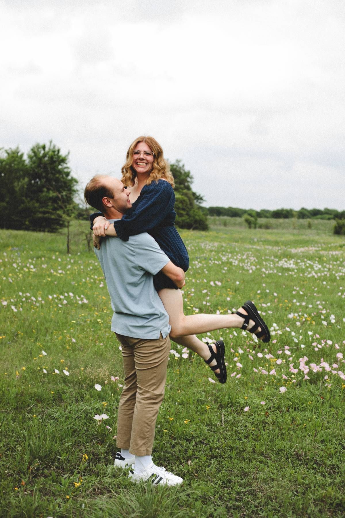 A smiling man lifts a laughing woman in his arms in a green wildflower meadow under an overcast sky.