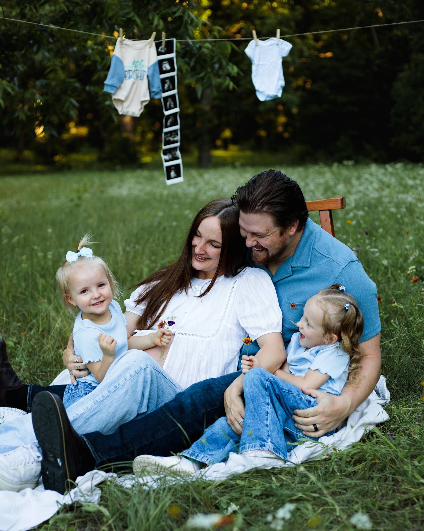 A smiling family of four with two young girls sits on a blanket in a green meadow beneath baby clothes hanging on a clothesline.