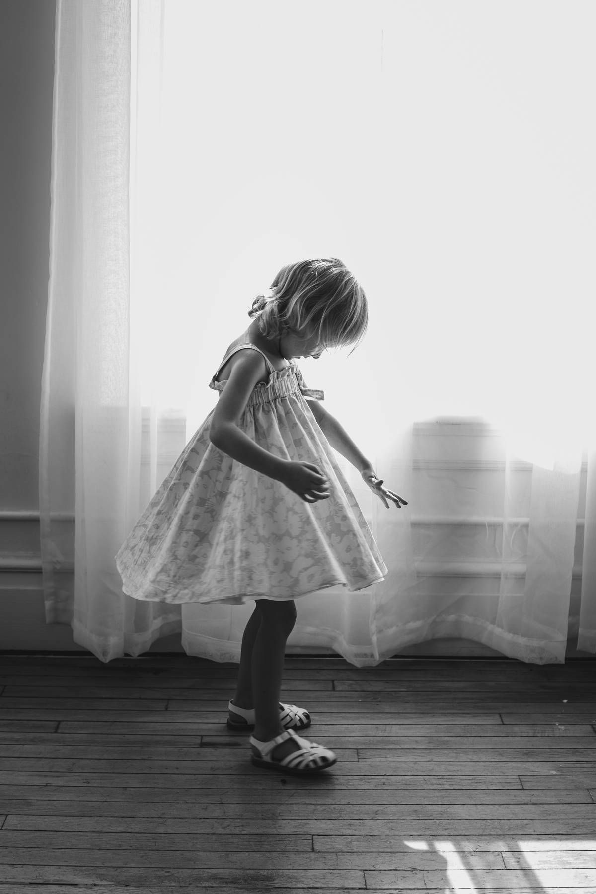 A black and white photo of a little girl in a floral dress twirling beside sheer curtains on a hardwood floor in soft window light.