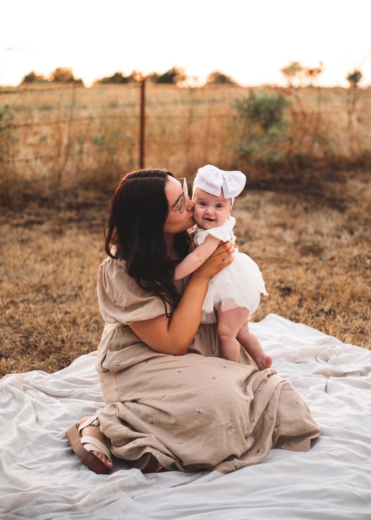 A mother in a neutral dress sits on a blanket in a golden field, kissing her baby girl wearing a white bow headband.
