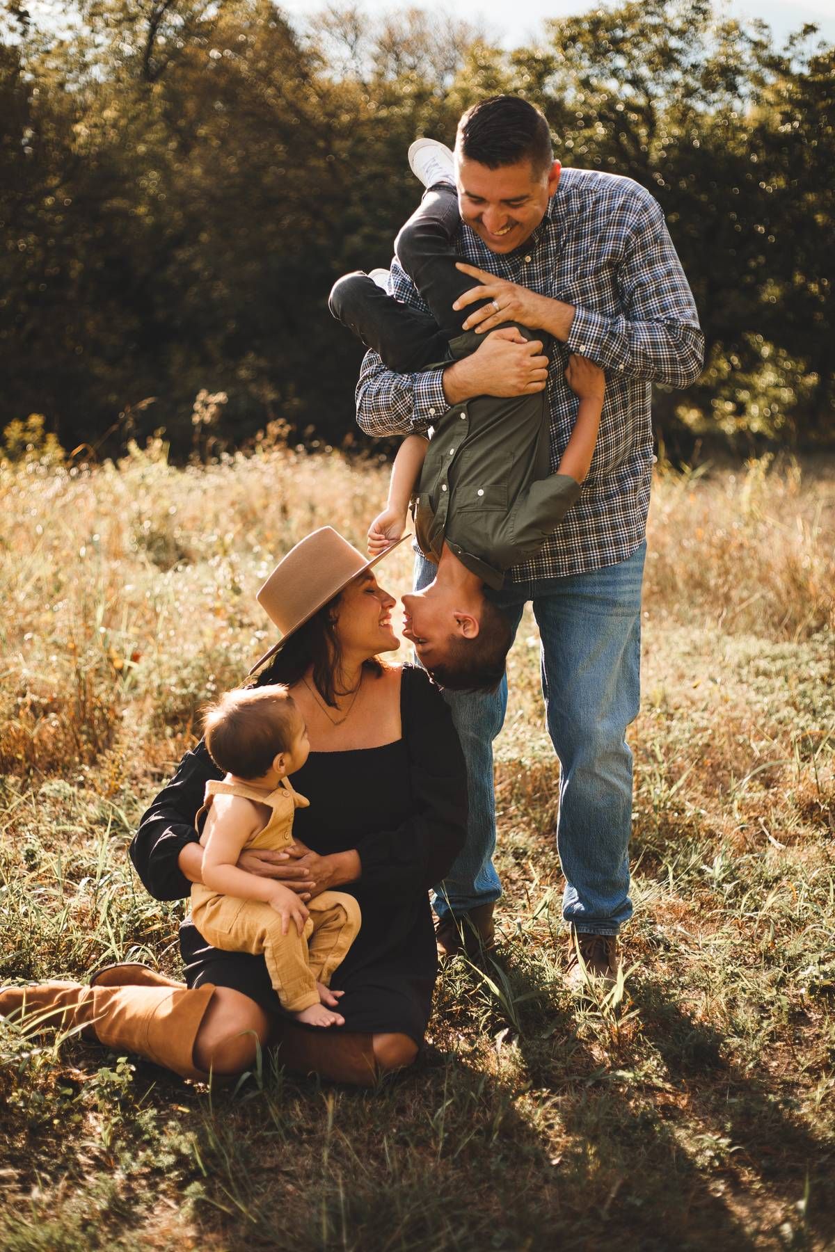 A father playfully flips his son upside down while a mother in a brown hat holds a baby on the grass in a sunlit field.