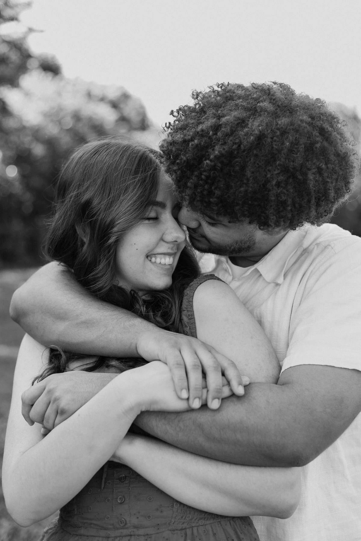 Black and white portrait of a smiling woman hugged tightly from behind by a curly-haired man kissing her cheek.