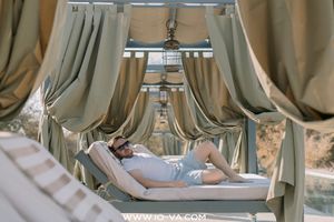 Person in sunglasses relaxes on a cushioned daybed inside a luxury poolside cabana with flowing olive-green curtains on a sunny day.