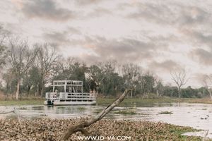 A white two-deck safari river boat floats on calm water beside a leafy shoreline under a soft, cloudy sky in an African wilderness.