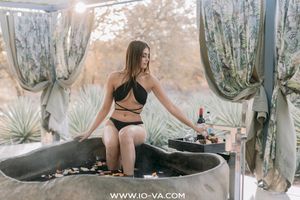 Woman in a black two-piece swimsuit sits on the edge of a rustic stone bathtub on an outdoor spa terrace with agave plants and drapes.