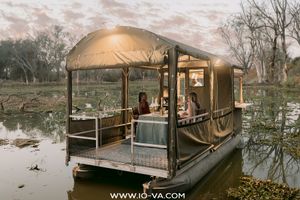 Two guests relax on a covered safari pontoon boat floating on calm, reflective river water surrounded by bare trees at sunset.