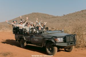 Group of smiling safari guests waving from an open-air game drive vehicle on a red dirt road with dry hills in the background.