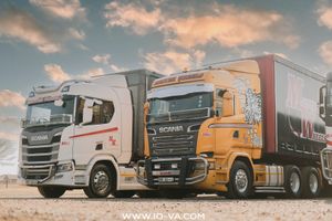 Two Scania semi-trucks, one white and one orange with a red trailer, parked side by side on a gravel lot under a dramatic cloudy sky.