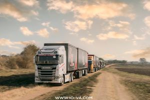 White semi-truck pulling a trailer along a dirt road through farmland, with a convoy of trucks behind under a cloudy sky.