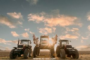 Three agricultural tractors parked side by side in an open field at sunset, with golden light shining through a dramatic cloudy sky.