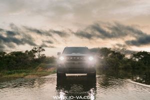 SUV with glowing headlights driving through shallow floodwater at sunset, with dramatic storm clouds and lush green trees in the background.