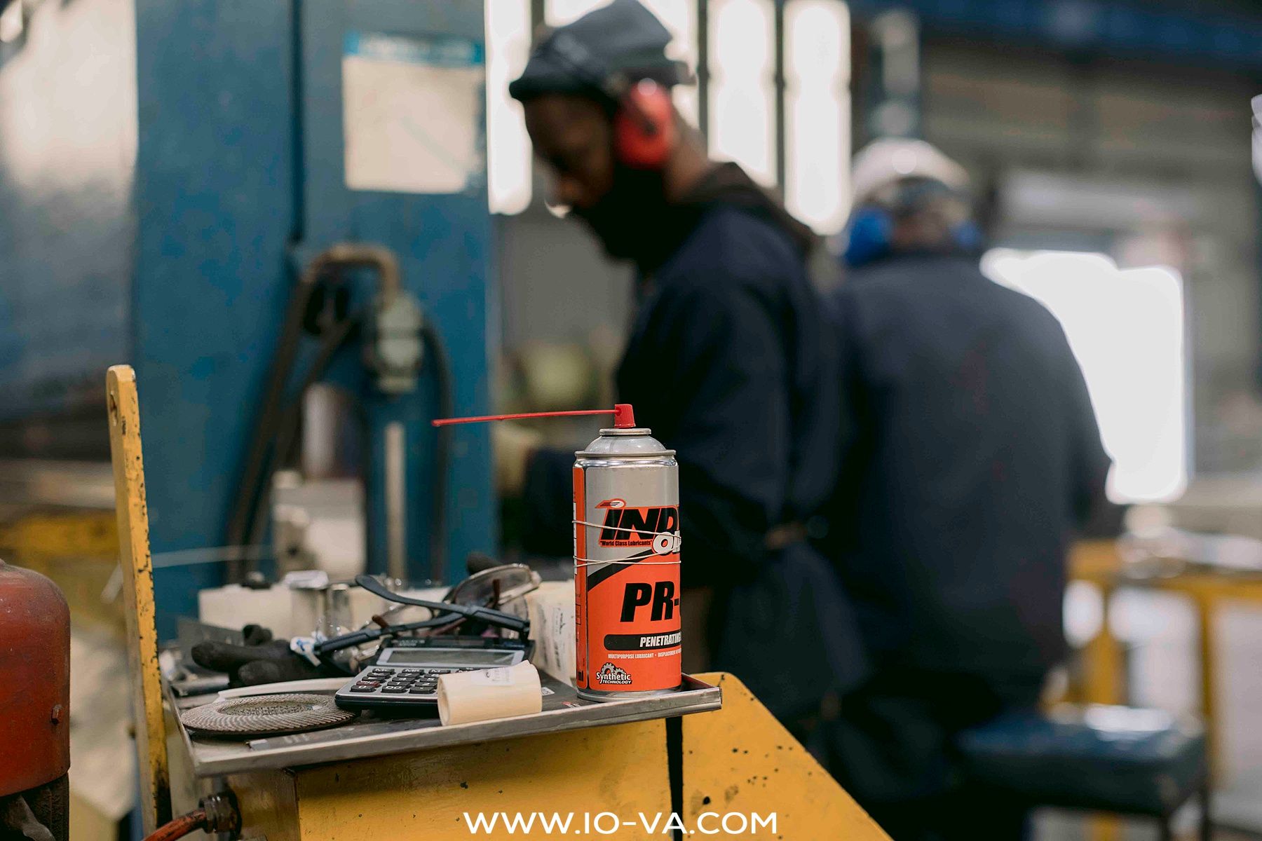 Workers in protective gear and ear protection stand in an industrial workshop behind a bench with tools and a spray can.