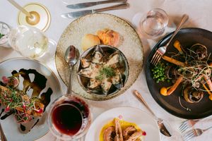 Overhead view of a restaurant table set with seafood dishes, roasted vegetables, a salad, red sauce, wine glasses and cutlery.