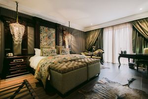 Luxurious hotel bedroom with a tufted ottoman, floral headboard, crystal chandelier, dark wood furniture, cowhide rug and drapes.