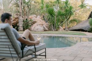 Man relaxing on a lounge chair holding a glass of wine beside a tranquil swimming pool surrounded by palm trees and natural boulders.
