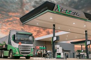 Green and white fuel tanker truck parked beside an X Fuels gas station with illuminated canopy and pumps under a dramatic orange sunset sky.