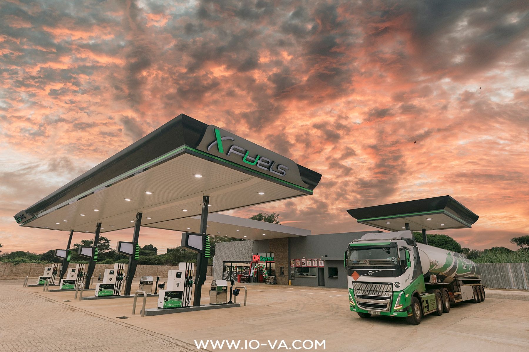 A green tanker truck parked beside a modern Xfuel gas station canopy with fuel pumps under a dramatic orange and purple sunset sky.
