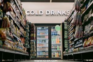 Convenience store aisle with shelves of snacks and a lit refrigerator display of bottled beverages under a "Cold Drinks" sign.